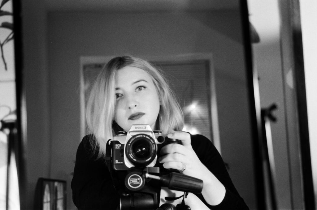 Elegant black and white portrait of a woman taking a selfie with an analogue camera in front of a mirror.