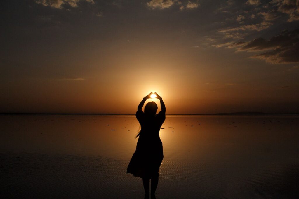 Silhouette of a woman with arms raised standing by the sea during a stunning sunset.